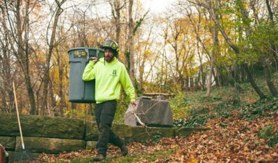 Independent Tree crew member in a neon hoodie and protective helmet carries a large gray bin filled with branches across a wooded backyard covered in fallen leaves, with a rake and blower nearby.