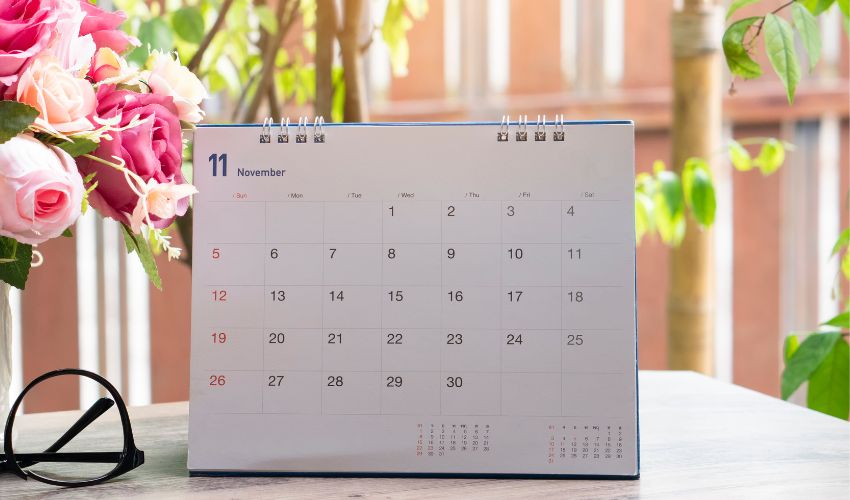 A desk calendar in front of a backyard with green trees and near a bouquet of cut flowers.