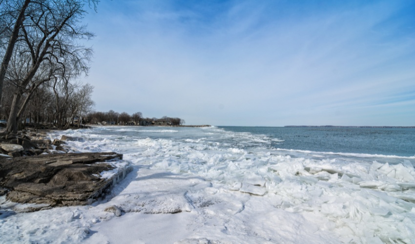 Lake Erie shoreline in winter with chunks of ice piled along the rocky shore, bare trees in background, and frozen lake water extending to the horizon under blue sky.