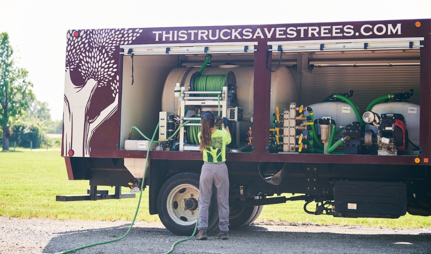 A plant health care technician in a safety vest stands at the rear of an Independent Tree treatment truck with specialized equipment and hoses visible.