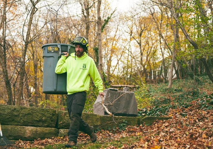 Independent Tree crew member in a neon hoodie and protective helmet carries a large gray bin filled with branches across a wooded backyard covered in fallen leaves, with a rake and blower nearby.